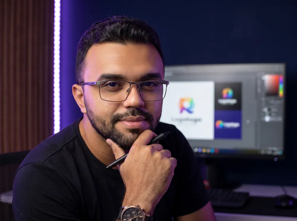 Diego Sued no estúdio, com a caneta da mesa digitalizadora apoiada no queixo e um logotipo aberto no monitor